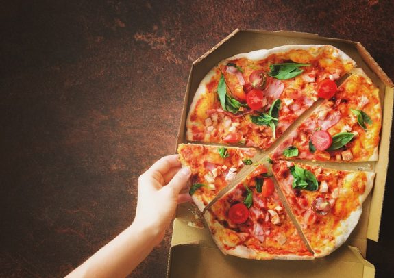 Female hand taking slice of fresh pizza from delivery box. Top view, dark background. Junk food. Sunny morning with sunlight. Toned pastel color