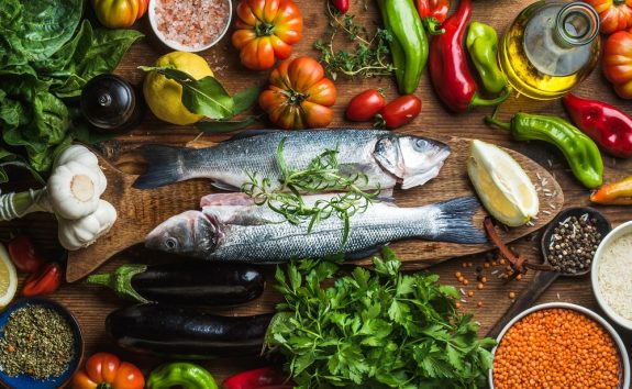 Raw uncooked seabass fish with vegetables, grains, herbs and spices on chopping board over rustic wooden background
